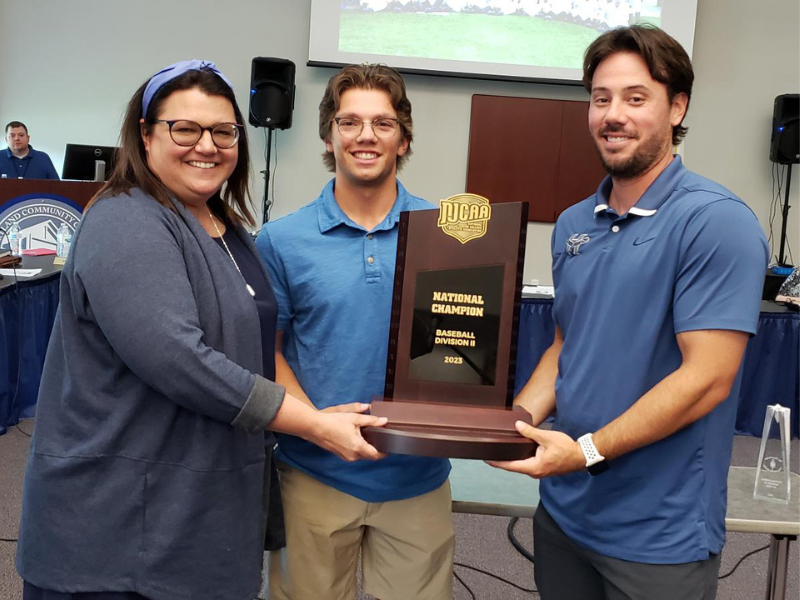Board member Becky Ropp, a baseball player, and baseball coach Chris Razzo holding the national championship trophey